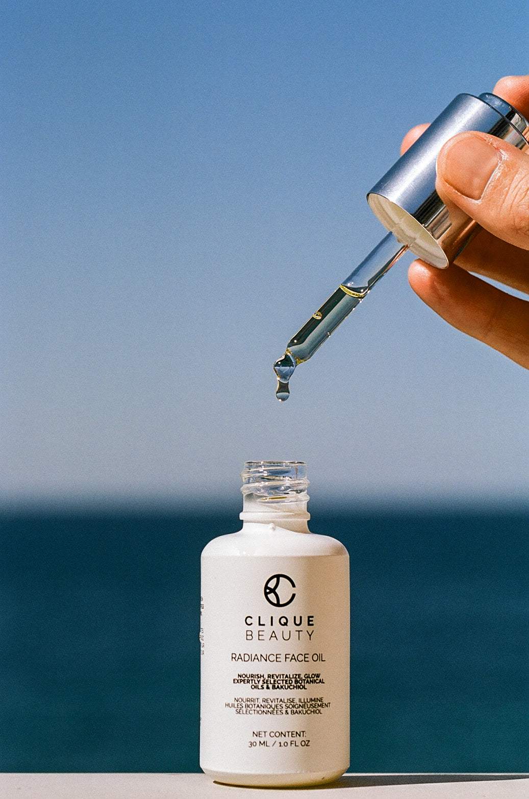 A hand holds a silver dropper above a white bottle of Clique Beauty Radiance Face Oil, with a drop of oil hovering over the open bottle against a blue sky and sea background.