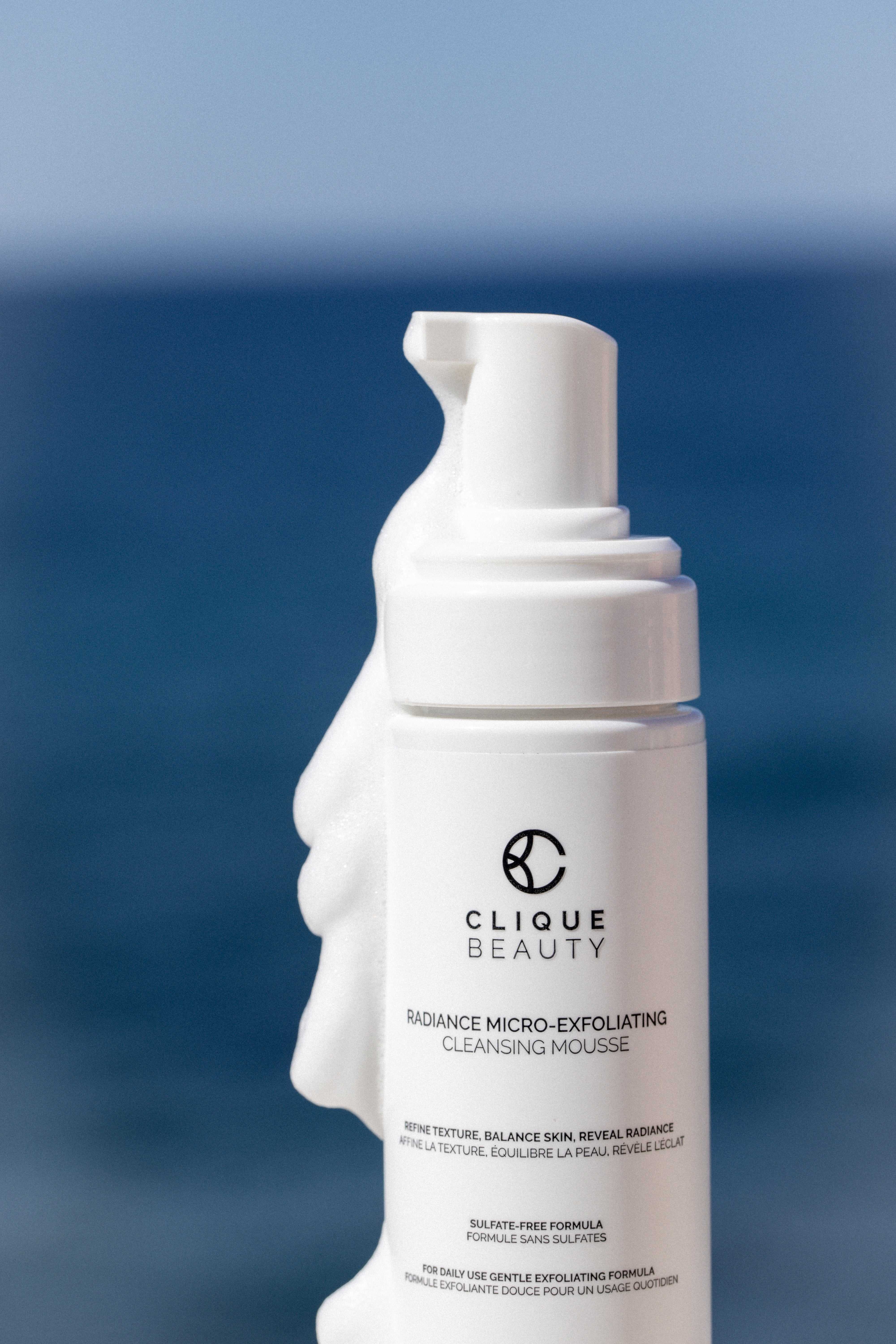 A white pump bottle of Clique Beauty Cleansing Mousse, held in front of a sculptural white hand, set against a blurred blue background.