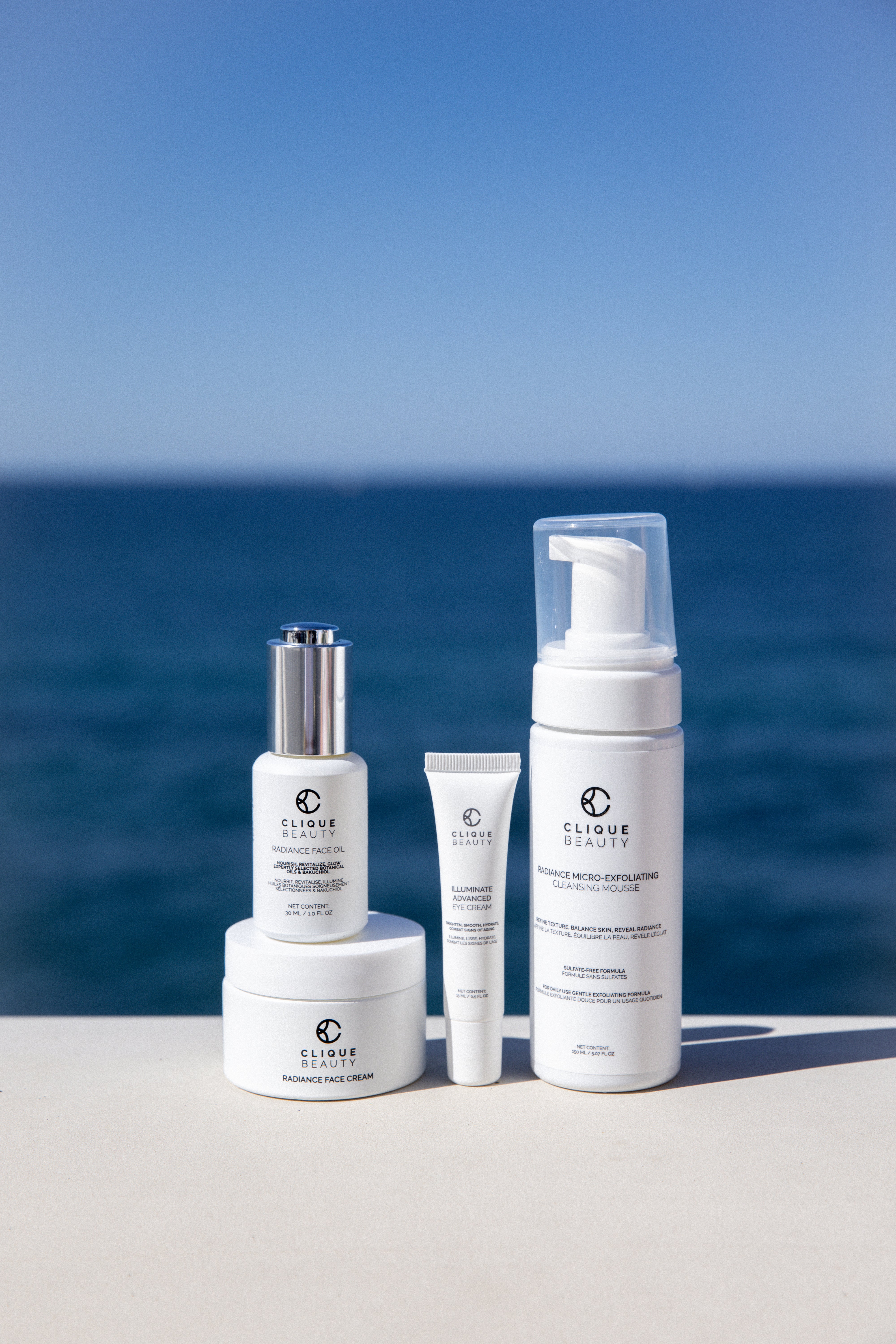 Four white skincare products from Clique Beauty Skincare arranged on a light beige surface with a scenic ocean and blue sky background. The set includes a stacked cream jar, a serum bottle with a silver cap, a small tube, and a larger pump bottle.