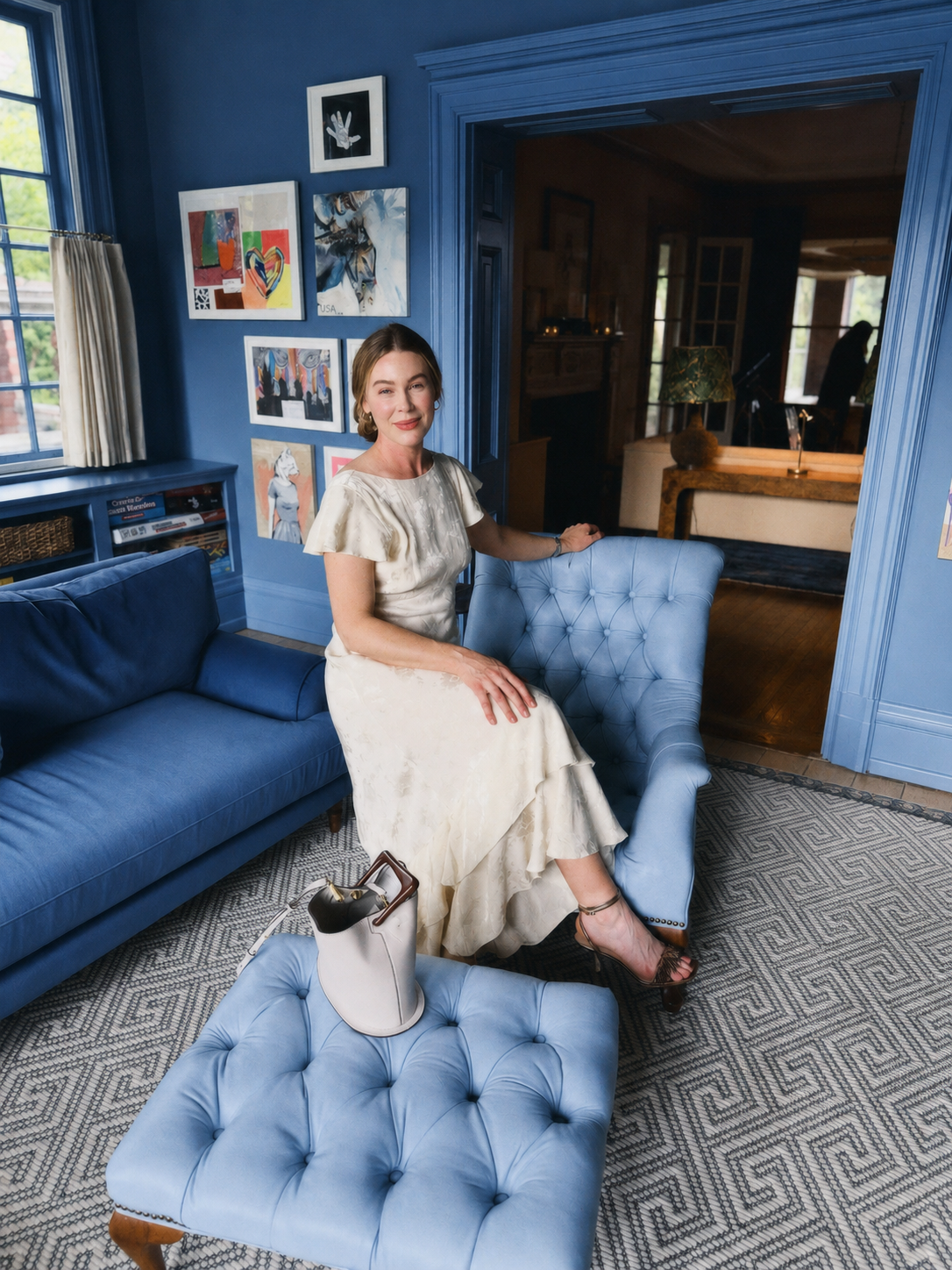 A woman in a cream ruffled dress sits on a tufted light blue armchair in a blue living room, with a matching blue sofa and patterned rug visible.