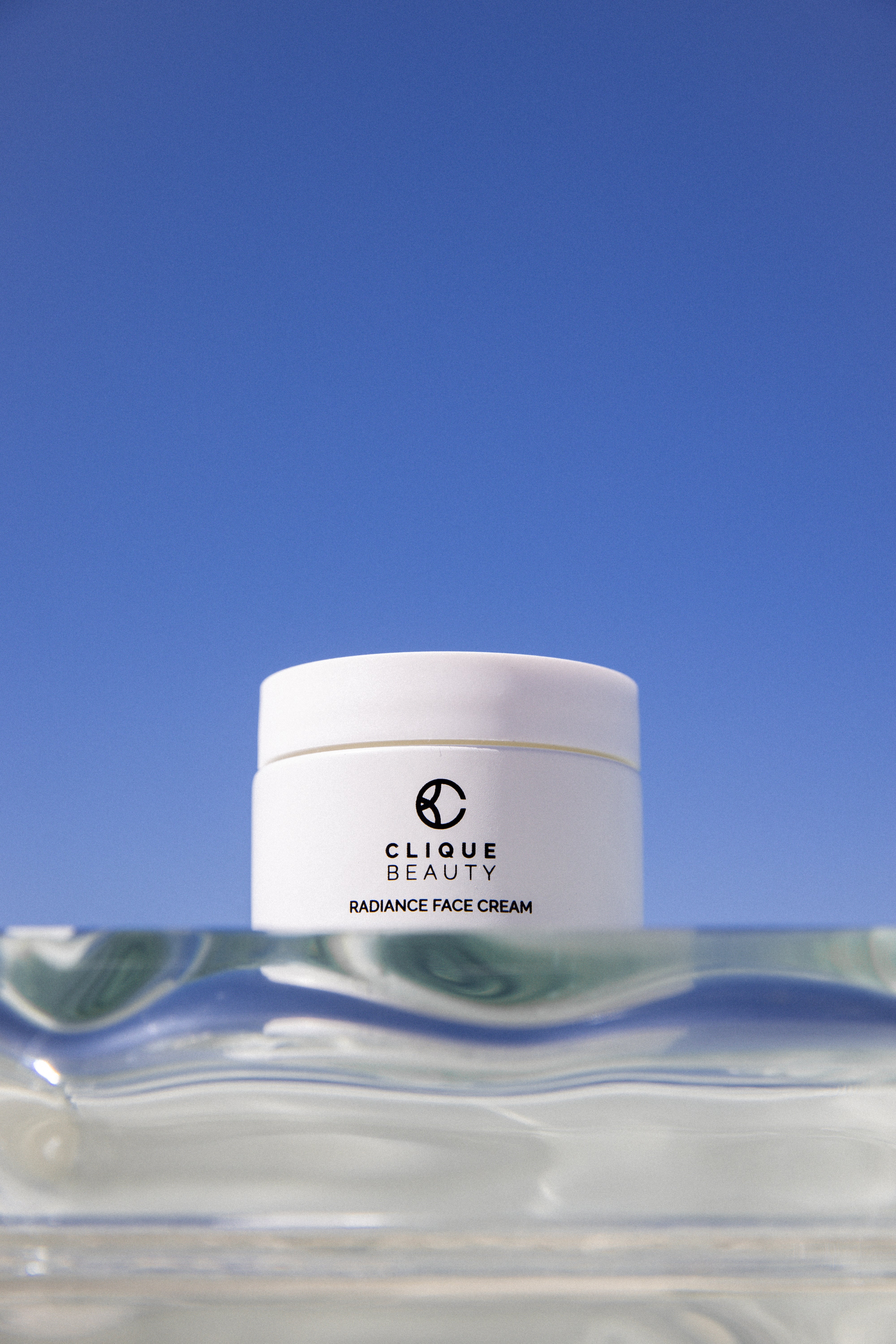 A white jar of Clique Beauty Radiance Face Cream sits on a clear glass surface against a solid blue sky background.