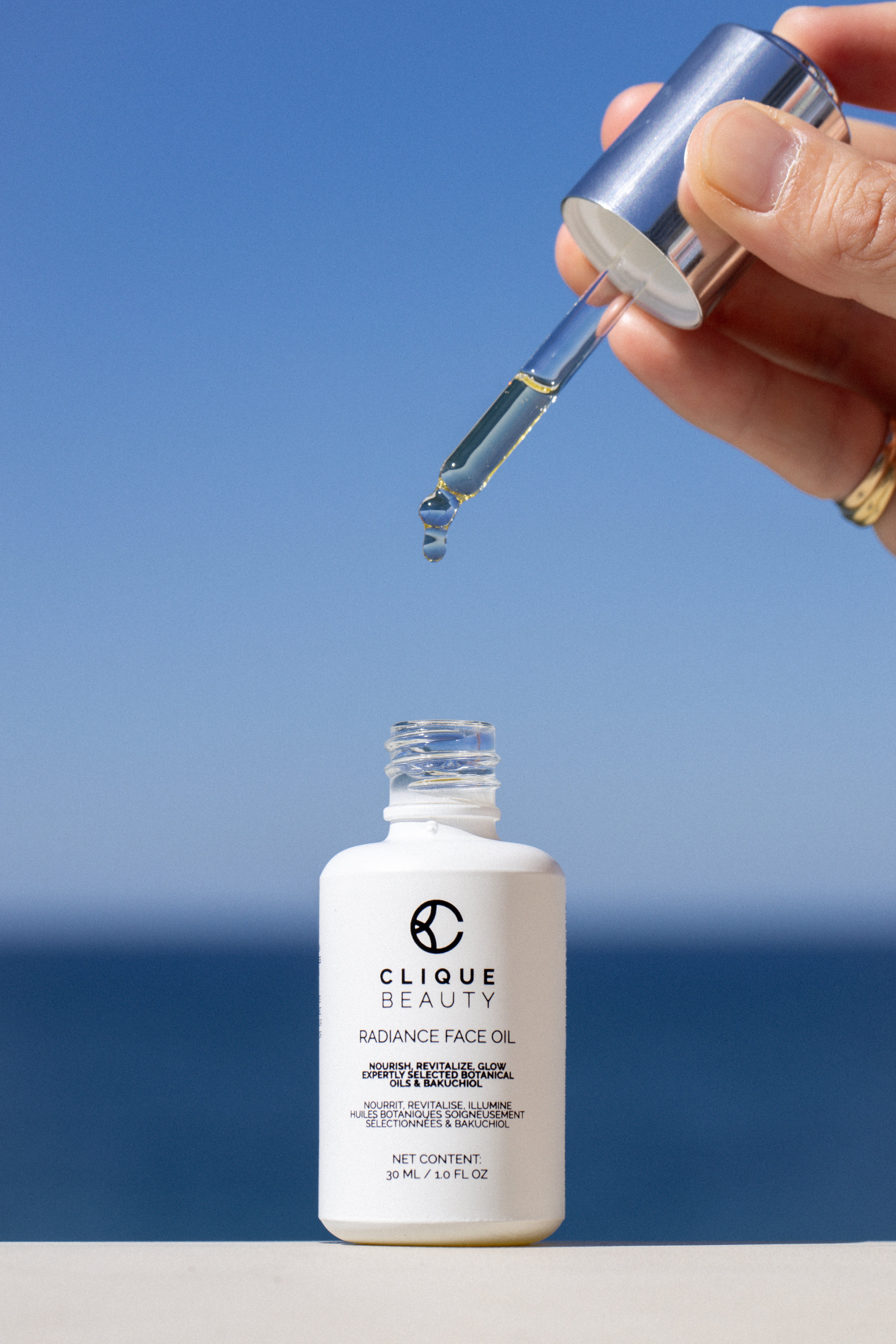 A hand holds a silver dropper above a white bottle of Clique Beauty Radiance Face Oil, with a clear glass dropper tip releasing a drop of oil against a blue sky and ocean background.