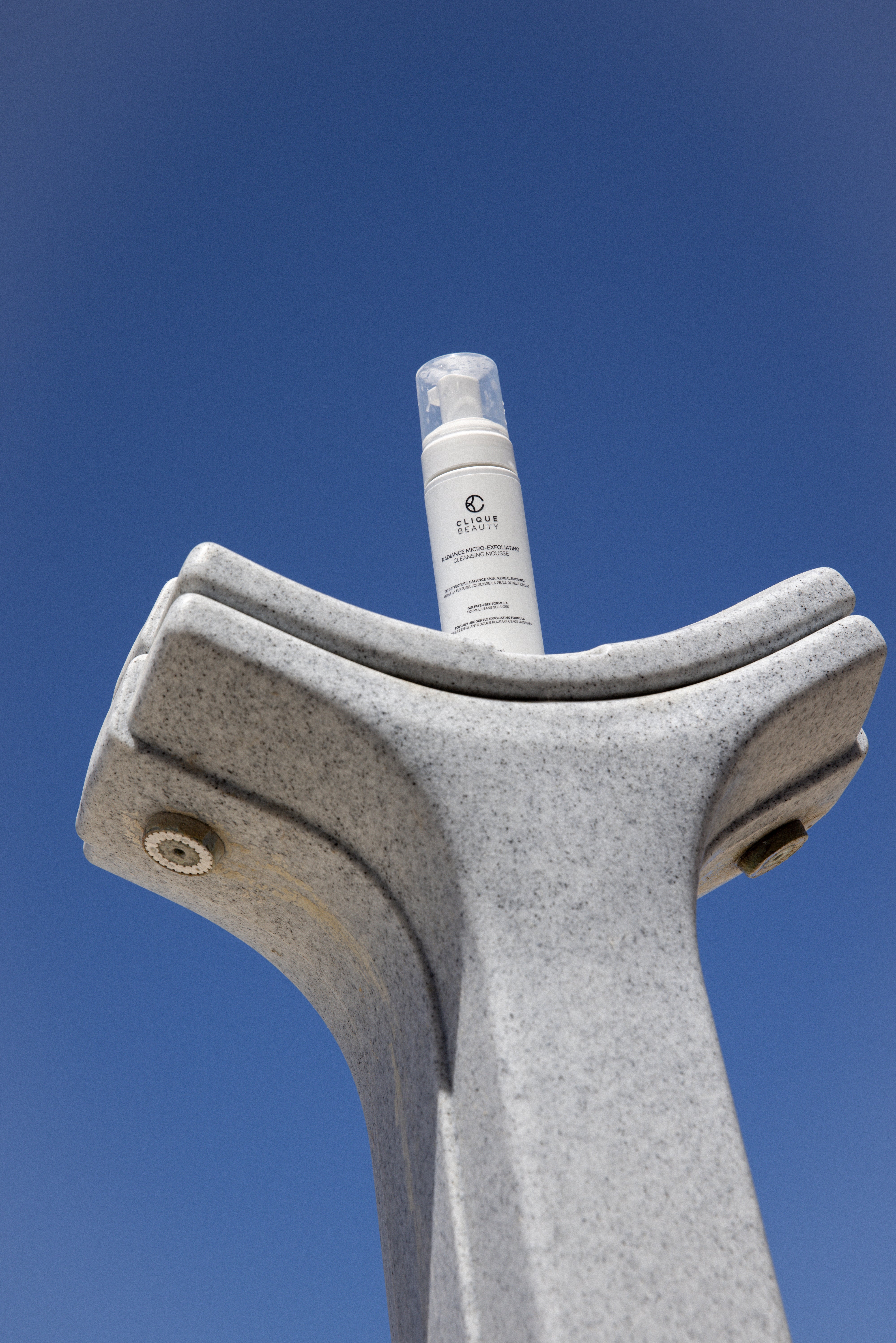 A white bottle of clique beauty, placed on top of a modern gray stone fountain against a clear blue sky.
